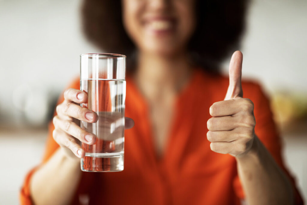 Frau mit gefülltem Wasserglas in der einen Hand, zeigt mit der anderen Hand den Daumen nach oben als Zeichen der Zustimmung für das Wasser