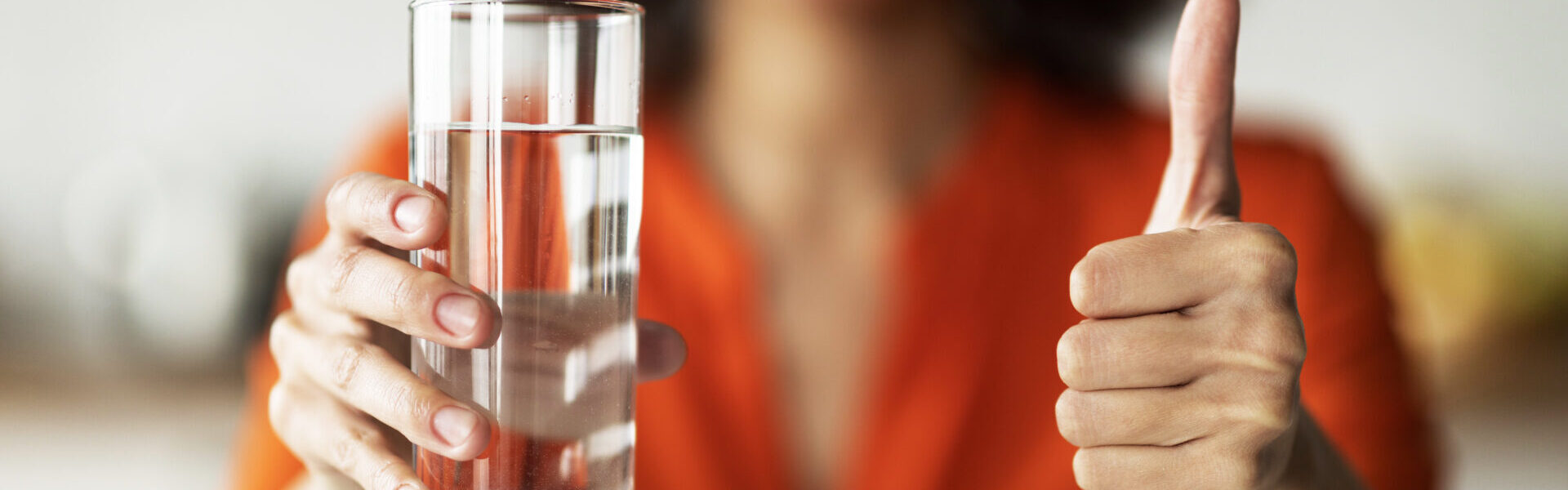 Frau mit gefülltem Wasserglas in der einen Hand, zeigt mit der anderen Hand den Daumen nach oben als Zeichen der Zustimmung für das Wasser.