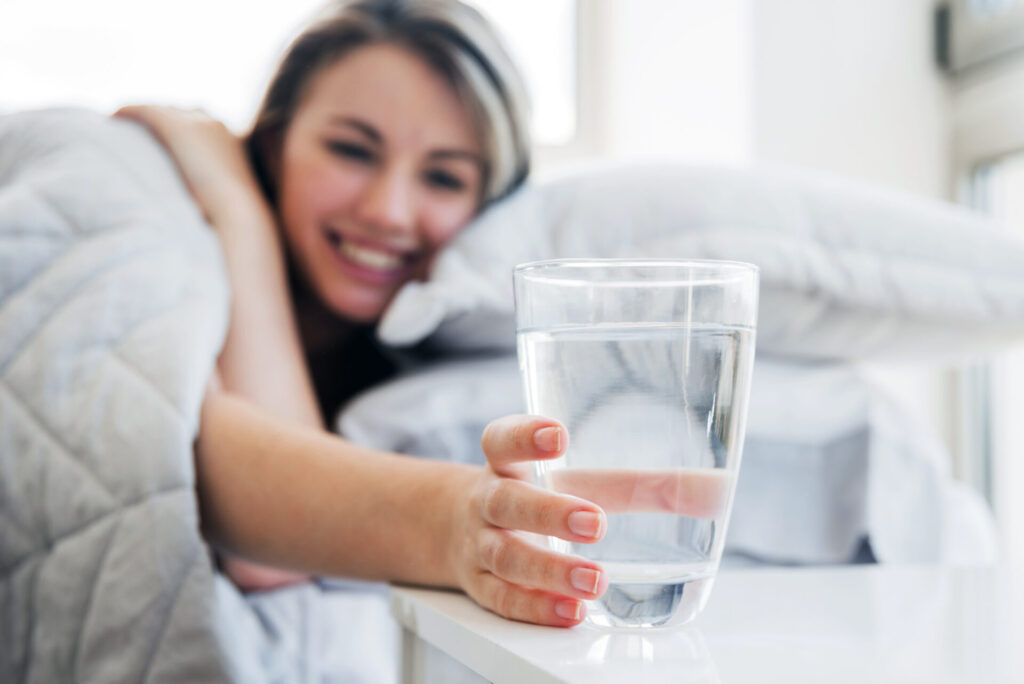 Frau im Bett greift nach Wasserglas auf Nachttisch.