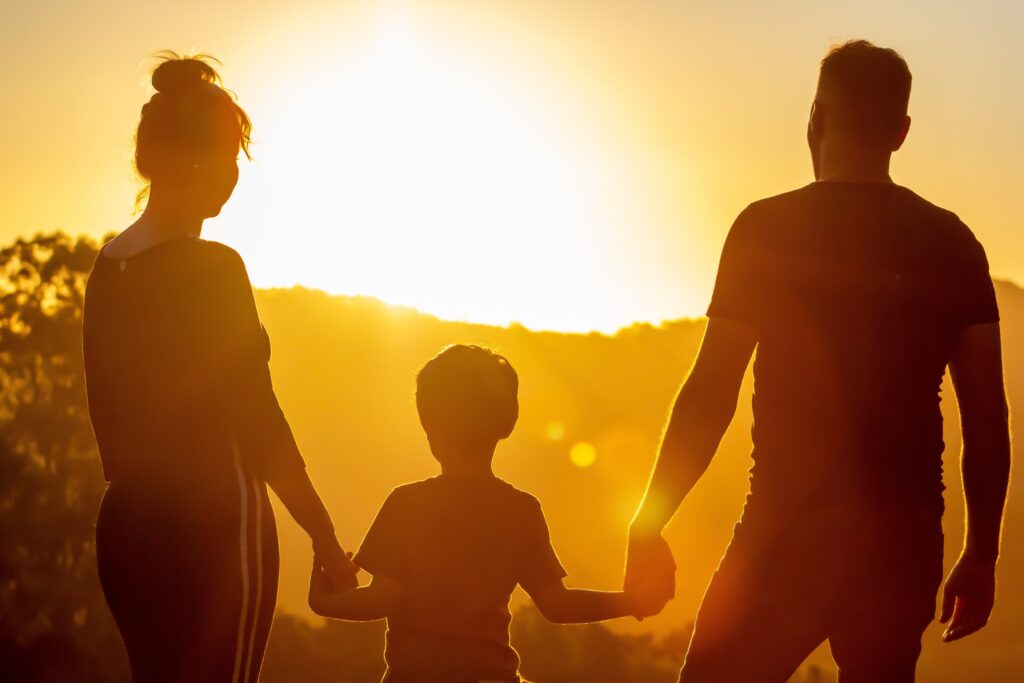 Stimmungsbild: Frau, Kind und Mann sind wie Schatten von hinten zu sehen, im Hintergrund geht die Sonne unter.