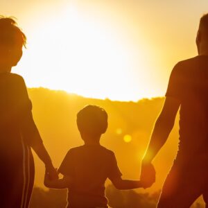 Stimmungsbild: Frau, Kind und Mann sind wie Schatten von hinten zu sehen, im Hintergrund geht die Sonne unter.