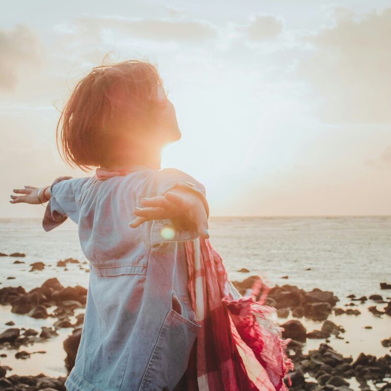 Kategoriebild: Frau mit ausgestreckten Armen geniesst die Sonne des Sonnenuntergangs, im Hintergrund Meer und Steinstrand.